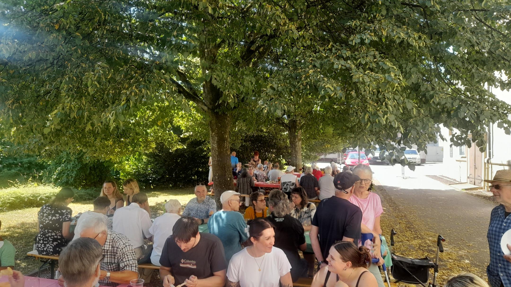 Unter den Bäumen sitzen Menschen  an Tischen, essen und unterhalten sich